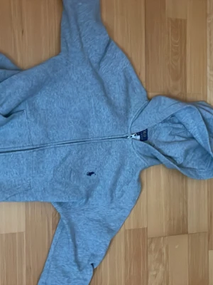 Grå zip-hoodie från Polo Ralph Lauren - Snygg gråmelerad hoodie med dragkedja från Polo Ralph Lauren. Klassisk huva, långa ärmar och fickor framtill. Liten mörkblå logga broderad på bröstet. Perfekt för en avslappnad och sportig stil.