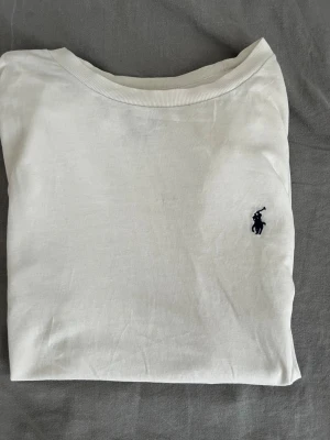 Vit t-shirt från Polo Ralph Lauren - Klassisk vit t-shirt från Polo Ralph Lauren med den ikoniska mörkblå loggan broderad på bröstet. Nypris kostar den runt 1000kr.  T-shirten har rund halsringning och korta ärmar. Tillverkad i mjuk bomull för en skön känsla och enkel stil. Den har även en qrkod som bevisar att den är äkta. Priset kan diskuteras eftersom de finns slitage som exempelvis svett märken men de syns inte alls när den används. 