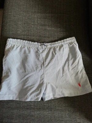 Randiga shorts med snörning - Snygga shorts med smala blå och vita ränder och elastisk midja med snörning. Shortsen har en liten röd broderad logga framtill och en bakficka med knapp. Perfekta för sommardagar och chill häng. Skriv privat för fler bilder eller om du har några frågor💗  