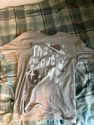 Beige t-shirt med planettryck - Snygg beige t-shirt med tryck av planeter och texten 'The Chaotic is Us'. T-shirten har rund hals och korta ärmar. Perfekt för dig som gillar unika och grafiska motiv.
