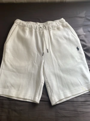 Vita shorts med snörning från Polo Ralph Lauren - Snygga vita shorts från Polo Ralph Lauren med elastisk midja och snörning. Klassisk logga broderad i mörkblått på vänster ben. Mjukt material och fickor på sidorna samt en bakficka. Perfekta för sommardagar och chill häng.