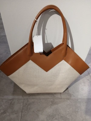 Beige och brun handväska - Stilren shoppingväska i beige canvas med bruna detaljer i konstläder. Väskan har två rejäla handtag, magnetknäppning och en innerficka med dragkedja. Perfekt för dig som vill ha plats för allt och ändå se trendig ut.