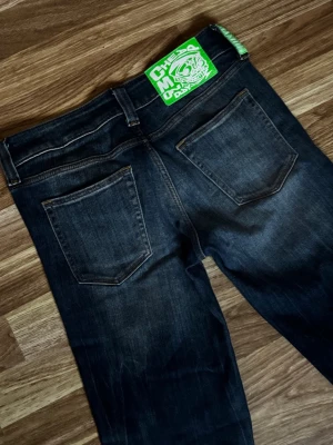 Mörkblå jeans från Cheap Monday - Säljer ett par mörkblå jeans med snygg slitning, lågmidjade och utsvängda ben från Cheap Monday. Jeansen har två bakfickor och en cool grön patch med tryck i midjan. Klassisk femficksmodell och kontrastsömmar. Perfekta för dig som gillar streetwear och vill sticka ut lite extra. Dom är i väldigt bra skick, knappt använda. Storlek 28/32. Pris går att diskutera💕💕