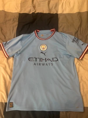 Manchester City matchtröja  - Snygg ljusblå Manchester City matchtröja från Puma med Haaland och nummer 9 på ryggen. Tröjan har klubbmärke, sponsorlogga och röda detaljer vid krage och ärmslut. Perfekt för dig som älskar fotboll och vill stötta City på matchdag!