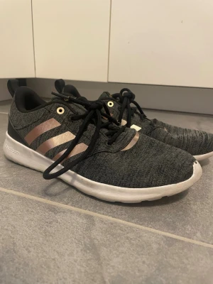 Adidas grå sneakers med kopparränder - Snygga Adidas sneakers i grått tyg med kopparfärgade ränder på sidorna och svart hälparti. Skorna har svarta snören, vit sula med cloudfoam-dämpning och en sportig, låg profil. Perfekta för en trendig och bekväm look.