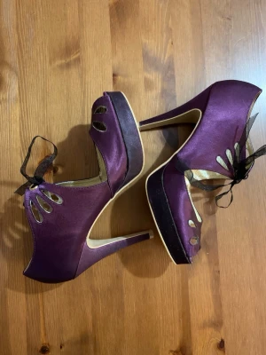 Lila satin-pumps med rosett från Bianco - Snygga lila pumps i satin från Bianco med peep-toe, utskurna detaljer och svart organzaband som knyts framtill. Skorna har hög klack och platåsula för extra höjd och stil. Perfekta för dig som vill sticka ut med en unik och elegant look.