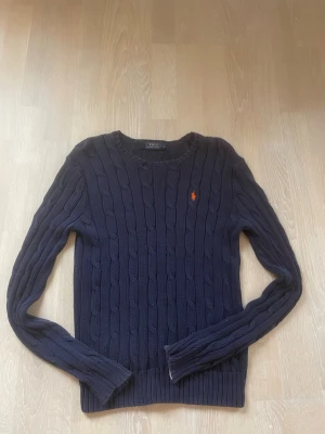 Mörkblå kabelstickad tröja Polo Ralph Lauren - Mörkblå kabelstickad tröja från Polo Ralph Lauren med rund halsringning och den klassiska röda logotypen broderad på bröstet. Tröjan har långa ärmar och ribbade muddar vid ärmslut och nederkant. Perfekt för lager på lager och stilren vardagslook.