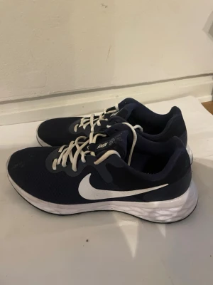 Blå Nike sneakers med vit sula - Snygga blå sneakers från Nike med vit sula och vit swoosh-logga på sidan. Skorna har vita snören och ovandel i meshmaterial för bra andningsförmåga. Perfekta för dig som gillar sportig stil och vill ha bekväma skor till vardags.