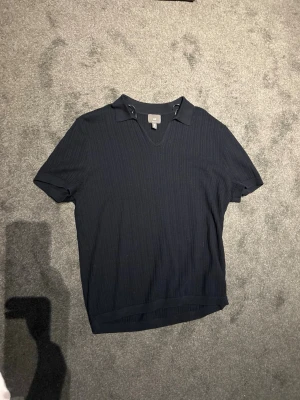 Mörkblå ribbad polotopp - Snygg mörkblå ribbad polotopp med kort ärm och v-ringning. Toppen har en klassisk krage och är tillverkad i ett mjukt material som känns skönt mot huden. Perfekt för en stilren och avslappnad look.