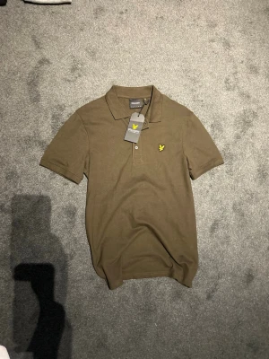 Olivgrön pikétröja från Lyle & Scott - Snygg olivgrön pikétröja från Lyle & Scott med klassisk krage och två knappar framtill. Tröjan har korta ärmar och den ikoniska gula örnloggan broderad på bröstet. Perfekt för en avslappnad men stilren look.