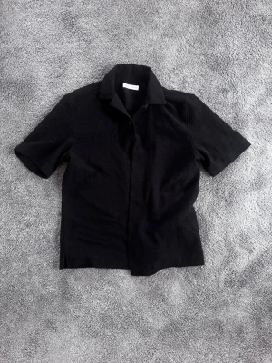 Svart kortärmad skjorta - Stilren svart skjorta med korta ärmar och klassisk krage. Skjortan har en rak passform och knäppning framtill. Perfekt för en enkel och clean look.