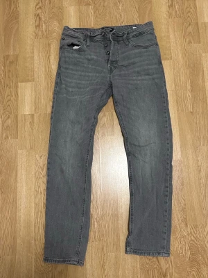 Grå raka jeans med knappgylf - Snygga grå jeans med raka ben och klassisk femficksmodell. Jeansen har knappgylf och är tillverkade i ett mjukt denimtyg som ger en avslappnad look. Perfekta för dig som gillar en enkel och stilren stil.