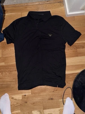 Svart Prada pikétröja med krage - Klassisk svart pikétröja med krage och knappar framtill. Tröjan har korta ärmar och en liten logodetalj på bröstet. Perfekt till jeans eller shorts för en clean och enkel stil. Storleken är mer s/m. Pris är inte spikat.