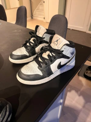 Svarta och vita Air Jordan 1 Mid - Säljer ett par Air Jordan 1 Mid sneakers i svart och vitt läder med klassisk Nike swoosh på sidan och Air Jordan-logga vid ankeln. Skorna har svarta skosnören och perforerad tå för extra ventilation. Perfekta för dig som gillar streetwear och snygg design.