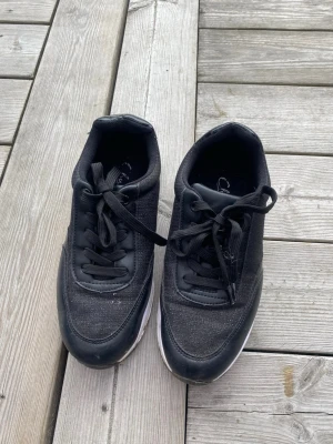Svarta sneakers med vit sula - Snygga svarta sneakers med vit sula och svarta skosnören. Ovandelen är i textil och syntetmaterial, vilket ger en sportig look. Passar perfekt till streetwear och vardagsoutfits. Klassisk låg modell som är enkel att matcha.
