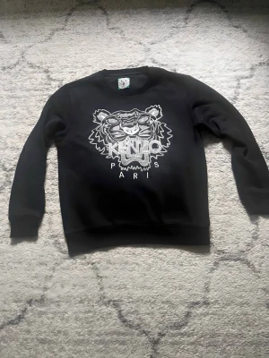 Svart Kenzo Tröja - Jag säljer en fin svart original kenzo tröja som är lite använd men fotfarande i bra skick som sagt den passar folk i längden 152-160 cm .För fler frågor det är bara o skriva.