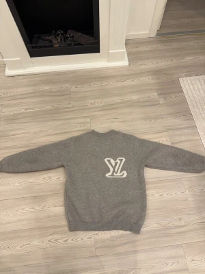 Grå tröja med LV-logga - Snygg grå tröja med stor vit LV-logga på ryggen. Tröjan har rund halsringning, långa ärmar och ribbade muddar vid ärmslut och nederkant. Perfekt för dig som gillar stilrena och exklusiva plagg.