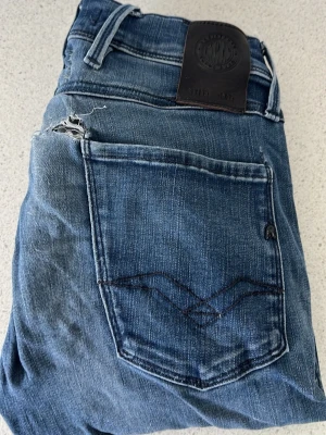 Replay anbass defekt  - Ett par klassiska blå jeans i denim med slitningar och hål vid bakfickorna. Jeansen har dubbla bälteshällor och en mörk läderpatch i midjan. Snygga detaljer på bakfickorna med mörk söm. Perfekta för en avslappnad och trendig look. Det är ett par defekt jeans med hål bak vid fickorna går att sy ihop W27 L32 