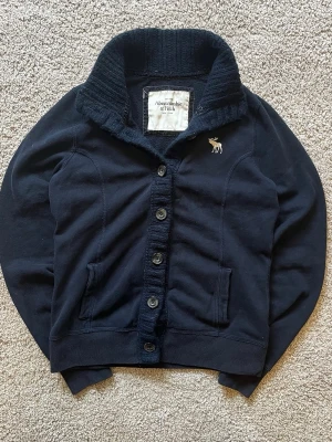 Mörkblå kofta från Abercrombie & Fitch - Mysig mörkblå kofta från Abercrombie & Fitch med grovstickad krage, knappstängning och två fickor framtill. Liten broderad älglogga på bröstet ger en klassisk touch. Perfekt för lager på lager och chill stil. Storlek L men sitter mer som S-M.
