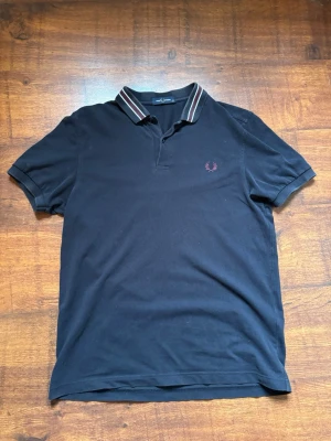 Marinblå pikétröja från fred Perry  - Klassisk marinblå pikétröja med korta ärmar och krage med vita och röda ränder. Tröjan har en liten broderad logga på bröstet och är tillverkad i bomull. Perfekt till en avslappnad och stilren look.