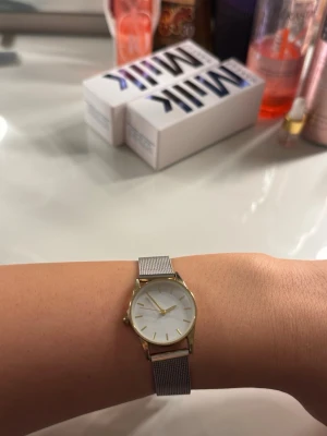Stilren silverfärgad meshklocka - Säljer en elegant klocka med silverfärgat mesharmband och rund vit urtavla. Klockan har guldfärgade detaljer på boetten och visarna, vilket ger en klassisk och stilren look. Perfekt accessoar för dig som gillar minimalistisk design.