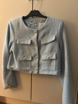 Ljusblå tweedkavaj med guldknappar - Trendig ljusblå kavaj i tweed med fyra fickor framtill och snygga silverknappar. Kortare modell med rundad halsringning och långa ärmar. Har fickor med öppning! Använd bara 1 gång