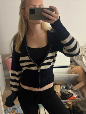 Marinblå randig zip-kofta - Trendig marinblå kofta med vita horisontella ränder och dragkedja framtill. Modellen är croppad med långa ärmar och ribbstickade muddar. Perfekt att slänga över ett linne för en cool och avslappnad look.
