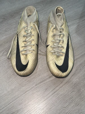 Nike Air Zoom Mercurial Superfly 9 - Säljer ett par beige fotbollsskor från Nike med stor svart swoosh på ovansidan. Skorna har snörning och låg profil, tillverkade i syntetmaterial för bra bollkontroll. Perfekta för dig som vill sticka ut på planen.