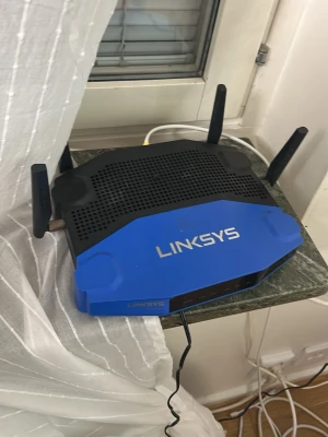  Linksys router Gaming - Säljer en router från Linksys i blått och svart med tre antenner. Perfekt för att förbättra wifi-täckningen hemma. Routern har en modern design med tydlig logga på ovansidan och flera portar på baksidan för nätverkskablar.