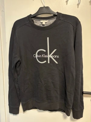 Svart sweatshirt från Calvin Klein - Stilren svart sweatshirt från Calvin Klein Jeans med stort vitt ck-tryck på bröstet. Tröjan har rund halsringning, långa ärmar och ribbade muddar vid ärmslut och nederkant. Perfekt för en avslappnad och trendig look.