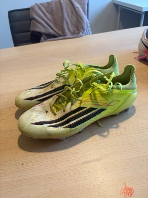 Gula fotbollsskor med svarta ränder - Säljer ett par snabba fotbollsskor i gul och vit färg med svarta ränder på sidan. Skorna har snörning och dobbar för bra grepp på planen. Lätta och smidiga, perfekta för dig som vill ha kontroll och fart på fotbollsplanen.