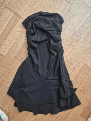 Svart axelbandslös klänning Shein - Snygg svart axelbandslös klänning från Shein i kort modell. Klänningen har rynkade detaljer på sidorna och en lätt utsvängd kjol. Perfekt för dig som vill ha en enkel men stilren look.