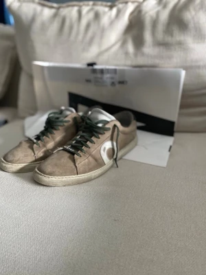 Oliver Cabell - Säljer ett par beige sneakers i mocka med gröna skosnören och vit sula. Skorna har en stor vit cirkeldetalj på sidan och mörkblå detalj vid hälen. Klassisk låg modell som passar till det mesta.