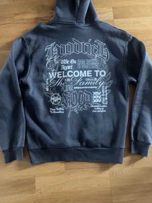 Grå hoodie med tryck från Hoodrich - Snygg grå hoodie från Hoodrich med stor vit text och grafiskt tryck på ryggen samt mindre tryck på bröstet. Klassisk känguruficka framtill och huva. Perfekt för dig som gillar streetwear och vill sticka ut med cool design.