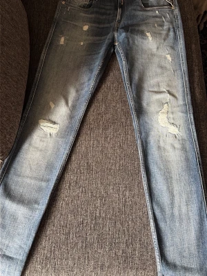 Ljusblå slitna jeans från Replay - Säljer ett par ljusblå jeans från Replay med slitningar och hål på benen för en trendig, avslappnad look. Jeansen har fem fickor, normal midja och smal passform. Perfekta för dig som gillar en casual och modern stil.
