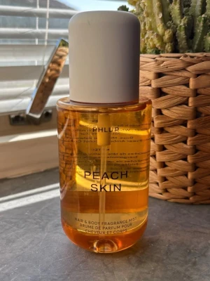 Peach Skin phlur  - Fräsch hår- och kroppsmist från Phlur med doften Peach Skin. Flaskan är cylinderformad i genomskinlig orange plast med vit rund kork. Perfekt för dig som vill ha en lätt och fruktig doft till både hår och kropp. 240ml. Testad 