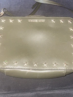 Olivgrön handväska med stjärnnitar - Snygg olivgrön handväska från Michael Kors i slätt läder. Väskan har silverfärgade stjärnnitar runt kanterna och en avtagbar axelrem. Perfekt för dig som gillar detaljer och vill sticka ut lite extra.