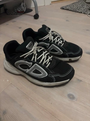 Dior B30 - Säljer ett par svarta Dior sneakers med tydliga silverdetaljer och vita snören. Skorna har en chunky sula och en modern, sportig design med mesh och läderpaneler. Perfekta för dig som vill ha en statement sneaker med exklusiv känsla.