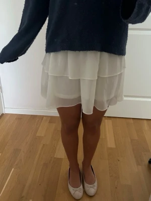 Vit volangkjol med smock - Söt vit kortkjol med bred smock i midjan och dubbla volanger. Kjolen har en luftig och flowy känsla, perfekt för varma dagar. Tillverkad i ett tunt, lätt tyg som ger en romantisk look. 