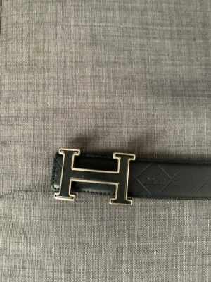 Svart bälte med H-spänne - Snyggt svart bälte med ett stort H-format metallspänne. Bältet har diskret rutmönster längs remmen och är gjort i skinn. Perfekt för att lyfta vilken outfit som helst.