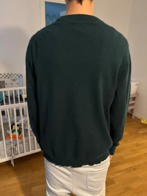 Mörkgrön stickad tröja med emblem - Mörkgrön stickad tröja med rund hals och ribbade muddar. På bröstet sitter ett litet broderat emblem i rött, vitt och guld. Tröjan har en klassisk passform och är perfekt för lager-på-lager. Materialet ser mjukt och bekvämt ut.