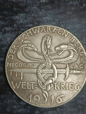En antik silvermedalj från WWI (första världskriget) - Ena sidan av medaljen har tre ormar och texten 'DER SCHWARZEN BERGE 1916', den andra visar tre kvinnor i antik stil med texten 'NIKITAS SCHICKSALS TOECHTER 1914'. Perfekt för samlare av historiska föremål från första världskriget!