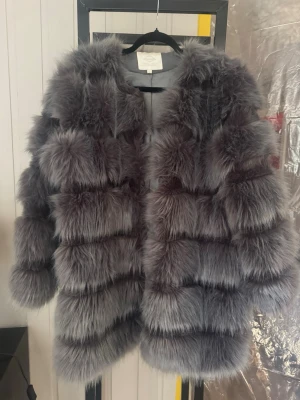 Grå fluffig pälsjacka - Säljer en superfluffig grå pälsjacka med breda horisontella paneler och rundad krage. Jackan har en oversized look och är gjord i fuskpäls som ger en lyxig och trendig vibe. Perfekt för att hålla värmen under kalla dagar och ger en statement-stil till din outfit.