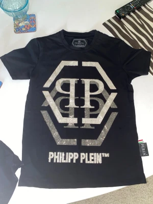 Svart Philipp Plein t-shirt med glitter - Snygg svart t-shirt från Philipp Plein med stort glittrigt logotryck framtill i silver. Klassisk passform och korta ärmar. Tillverkad i mjuk bomull för skön känsla. Perfekt för dig som gillar statement-plagg och exklusiv streetstyle.