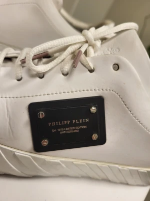 Philipp Plein Placca Nappa leather low-top sneakers, Storlek 39 - Snygga vita sneakers från Philipp Plein med exklusiv metallplatta på sidan och präglad logga. Skorna har vita snören och en unik sula med räfflat mönster. Tillverkade i slätt läder för en clean och modern look.
