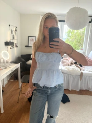 Ljusblå ärmlös linnetopp - Säljer en söt ljusblå linnetopp med smala axelband och volang nedtill. Toppen har en luftig passform och markerad midja, perfekt för varma dagar. Matcha med jeans eller kjol för en fräsch look.
