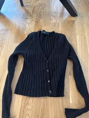 Svart ribbad kofta från Vero Moda - Snygg svart ribbad kofta från Vero Moda med v-ringning och knäppning framtill. Koftan har långa ärmar och smal passform, perfekt att styla över ett linne eller t-shirt. Tillverkad i mjukt material som känns skönt mot huden.