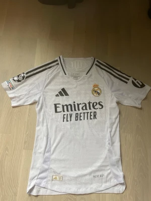 Real Madrid matchtröja Vini Jr #7 - Säljer en vit Real Madrid matchtröja från Adidas med Vini Jr och nummer 7 på ryggen. Tröjan har svarta detaljer, klubbmärke på bröstet och sponsortryck. Tillverkad i lätt funktionsmaterial med HEAT.RDY-teknologi och korta ärmar.