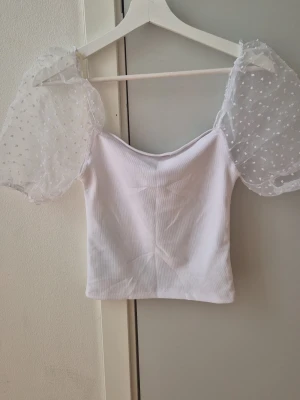 Vit topp med puffärmar och spetsdetaljer - Supersöt vit topp med transparenta puffärmar i spets och små prickar. Toppen har en croppad passform och ärmlös kropp med axelbandslös design. Perfekt för dig som vill sticka ut med en romantisk och trendig look. Storlek S