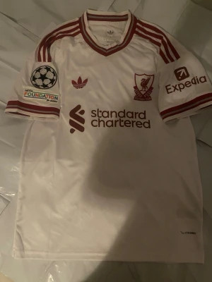 Vit Liverpool matchtröja Adidas - Säljer en vit Liverpool matchtröja från Adidas med röda detaljer och tryck. Tröjan har klubbmärke, sponsorloggor och Champions League-märke på ärmarna. Nummer 9 och namnet ISAK på ryggen. Tillverkad i lätt och ventilerande material.